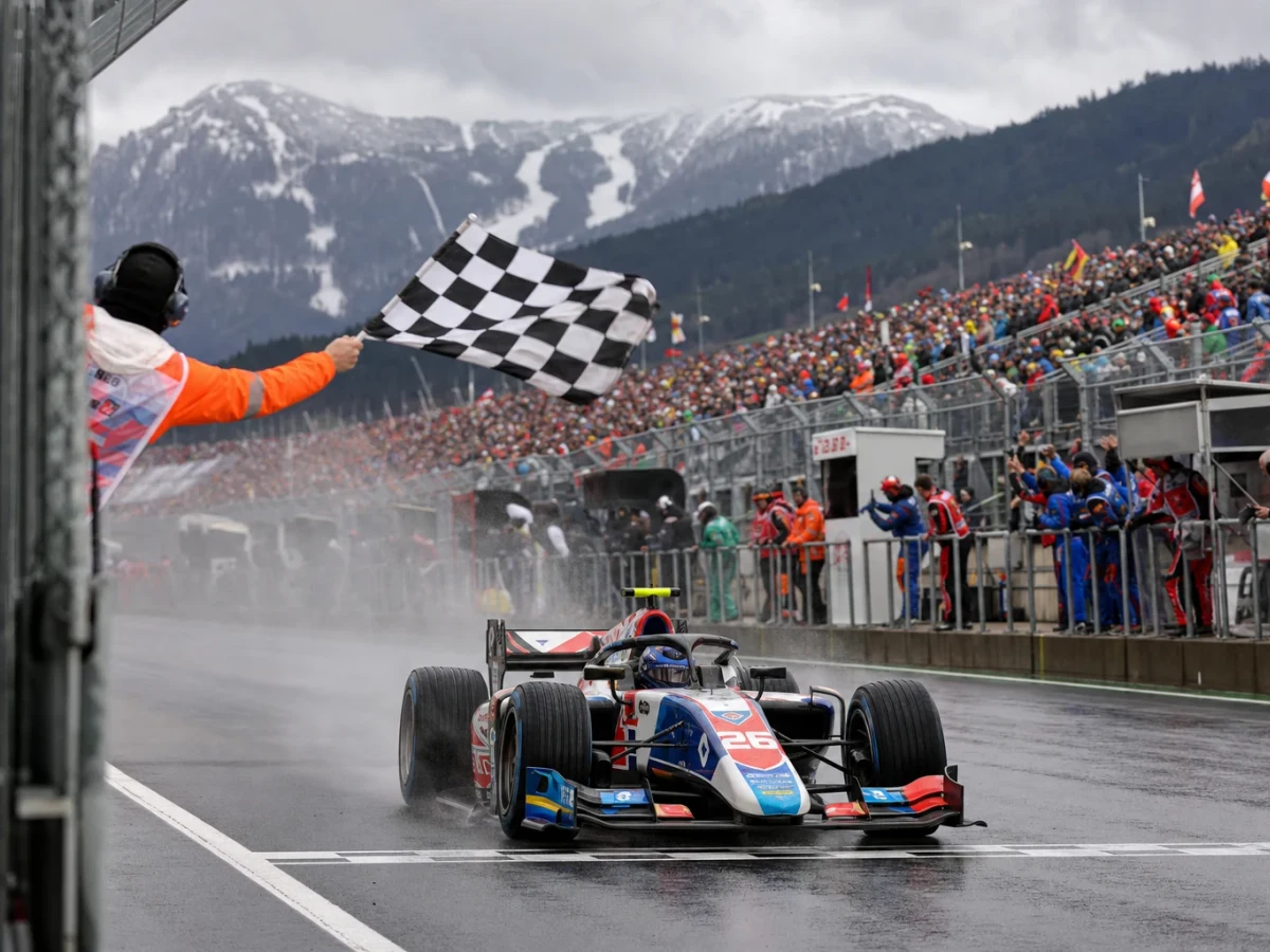 Silverfärgad formel 1-bil leder loppet över mållinjen på en bergig österrikisk racerbana i solljus.